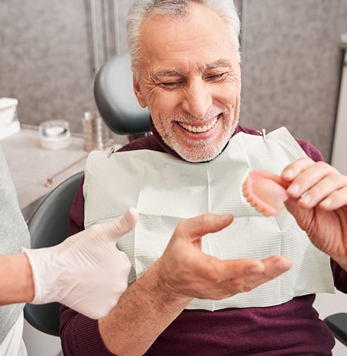 Man in dental chair smiling holding dentures with dentist’s gloved hand giving thumbs up